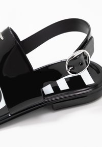 Sandales vernies noires avec une large partie supérieure, un design incurvé, une bride ajustable à la cheville et une boucle en argent. L'intérieur présente des rayures blanches contrastantes.
