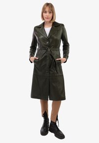 Trench-coat en cuir vert avec un design croisé, manches longues, deux poches avant et une ceinture à nouer à la taille. Texture lisse.