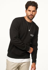 Black sweatshirt with long sleeves, ribbed cuffs, and crew neckline. Features small graphic detail on the chest. Layered over a white shirt.