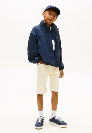 Niño de pie vistiendo una chaqueta azul marino, gorra a juego, pantalones cortos blancos, calcetas blancas y zapatillas azul marino contra un fondo liso.