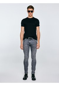 Noor mees mustade päikeseprillidega, musta t-särgi, hallide teksade, musta vöö ja mustade saapadega seisab ühevärvilise hele tausta ees.