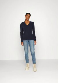 Pull en tricot marine à col en V avec manches longues, associé à un jean bleu clair et des bottines beiges à lacets, sur un fond uni.