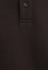 Brown polo shirt fabric up close, featuring textured ribbing and a black button near the collar. No visible logos or additional details.