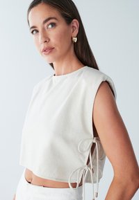 Hvid cropped top med rund hals og korte ærmer, der har bindebånd i siderne og et struktureret stof.