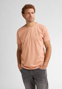 T-shirt a maniche corte color pesca chiaro in cotone, con scollo rotondo e un piccolo logo sul petto, abbinata a jeans grigi.