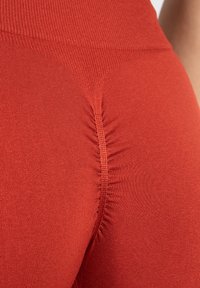 Leggings rossi realizzati in un tessuto testurizzato con cuciture arricciate sul retro, che evidenziano un design aderente e una superficie liscia.