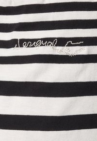 Mustekki ja valkoinen raidallinen kangas, jossa on tekstuuri hopeanvärinen brodeerattu logo ja hienovaraiset koristeommel-yksityiskohdat.