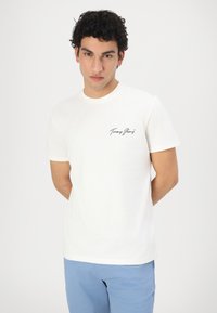 Camiseta de algodón blanca con cuello redondo, mangas cortas y logo negro de "Tommy Jeans" en la parte izquierda del pecho. Diseño sencillo y ligero.