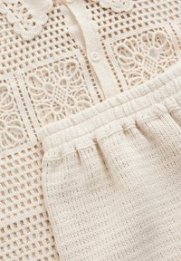 Tenue en tricot crème, comprenant un haut en dentelle crocheté texturé avec fermoir à boutons et une jupe ajustée en tricot côtelé.
