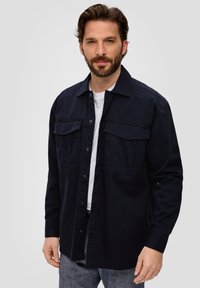Camicia botton-down di un blu scuro in cotone, con due tasche sul petto, maniche lunghe e taglio dritto. Indossata sopra una T-shirt bianca.