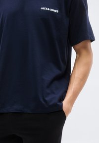 Άνδρας που φοράει μπλε σκούρο κοντομάνικο T-shirt Jack & Jones με το ένα χέρι στην τσέπη μαύρου παντελονιού, μπροστά σε απλό φόντο.