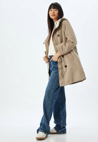 Abrigo beige con capucha, cierre de botones y cordón, combinado con una blusa de color claro y unos pantalones vaqueros azules sueltos. Zapatillas blancas completan el conjunto.
