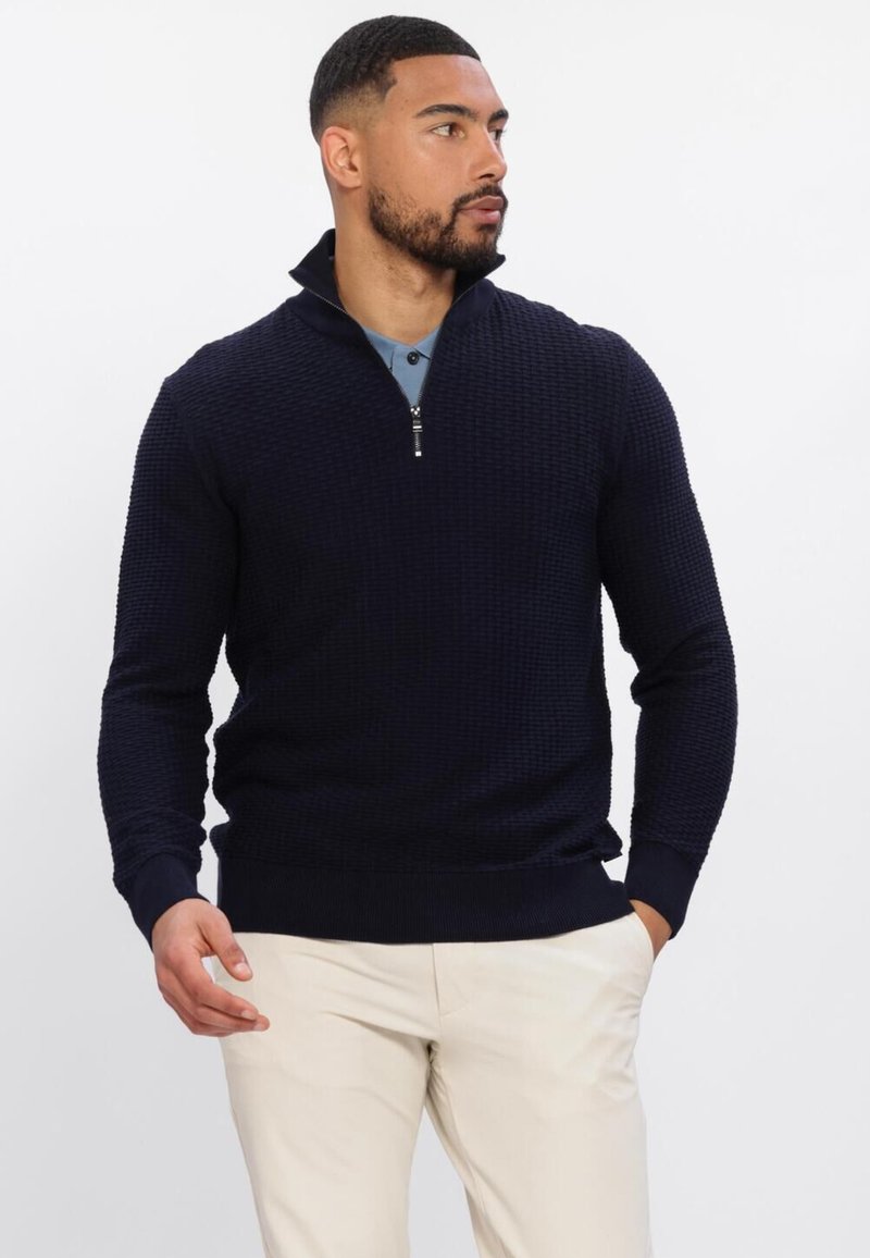 Homme portant un pull bleu marine texturé à demi-fermeture éclair sur une chemise bleue et un pantalon beige, regardant sur le côté sur fond blanc.