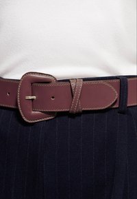 Bruine leren riem met een rechthoekige gesp, voorzien van contrasterende stiksels, gedragen over een donkere stof met krijtstreep.