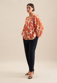 Blusa arancione e crema con motivo a fantasia, scollatura a V e maniche a palloncino, abbinata a pantaloni navy e sandali neri con tacco.