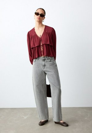 Mujer con gafas de sol, blusa burdeos de capas con lazos, vaqueros grises de talle alto y bailarinas marrón oscuro, de pie contra una pared blanca.