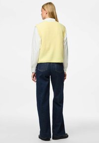 Majica bez rukava u žutoj boji, složena over bijele majice dugih rukava, u kombinaciji s tamnim trapericama širokih nogavica. Jednostavan, teksturiran dizajn.