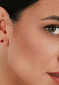 Rote Ohrstecker mit facettiertem Design, am Ohrläppchen positioniert. Die Hautstruktur zeigt einen natürlichen Look. Nahaufnahme des Gesichts und des Ohrarei.