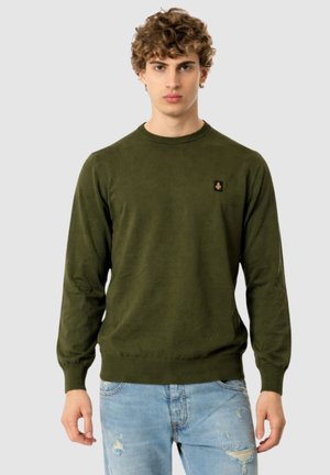 Giovane uomo con capelli ricci che indossa un maglione a maniche lunghe verde oliva e jeans chiari strappati, in piedi davanti a uno sfondo neutro.