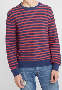 Gestreifter Pullover in Rot und Marineblau mit Rundhalsausschnitt, gerippten Bündchen und Saum. Der Stoff wirkt weich und hat eine gestrickte Textur. Wird über distressed Jeans getragen.