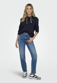 Felpa blu navy con lacci rosa e logo a forma di cuore, abbinata a jeans in denim blu e sneakers blu navy e bianche. Stile casual, vestibilità rilassata.