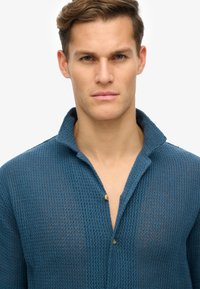 Blå struktureret strikskjorte med krave, der har knaplukning, letvægtsmateriale og åndbart design.