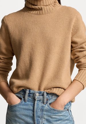 Person wearing a beige knitted turtleneck sweater tucked into light blue high-waisted jeans with hands in front pockets.
