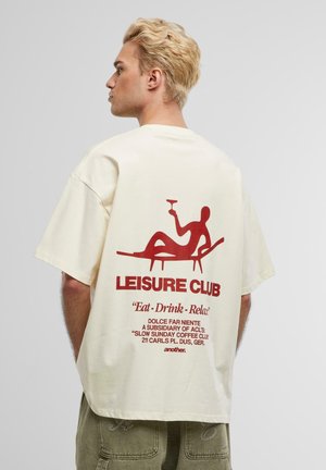 T-shirt crème à manches courtes avec un graphique rouge représentant une silhouette allongée tenant un verre, avec le texte "LEISURE CLUB" au-dessus et des détails supplémentaires de l'offre en dessous.