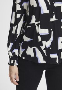 Blusa nera con motivo astratto bianco e lavanda, con chiusura a bottoni e polsini arricciati. Il materiale sembra leggero e morbido.