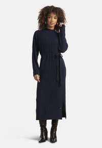 Vestito lungo a maglia blu navy con motivo testurizzato, maniche lunghe, vita con cintura e spacco laterale. Abbinato a stivali neri al ginocchio.