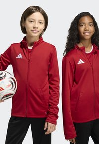 Deux jeunes en vestes de sport rouges se tiennent côte à côte, l'un tenant un ballon de football blanc et noir avec le texte "TIRO".