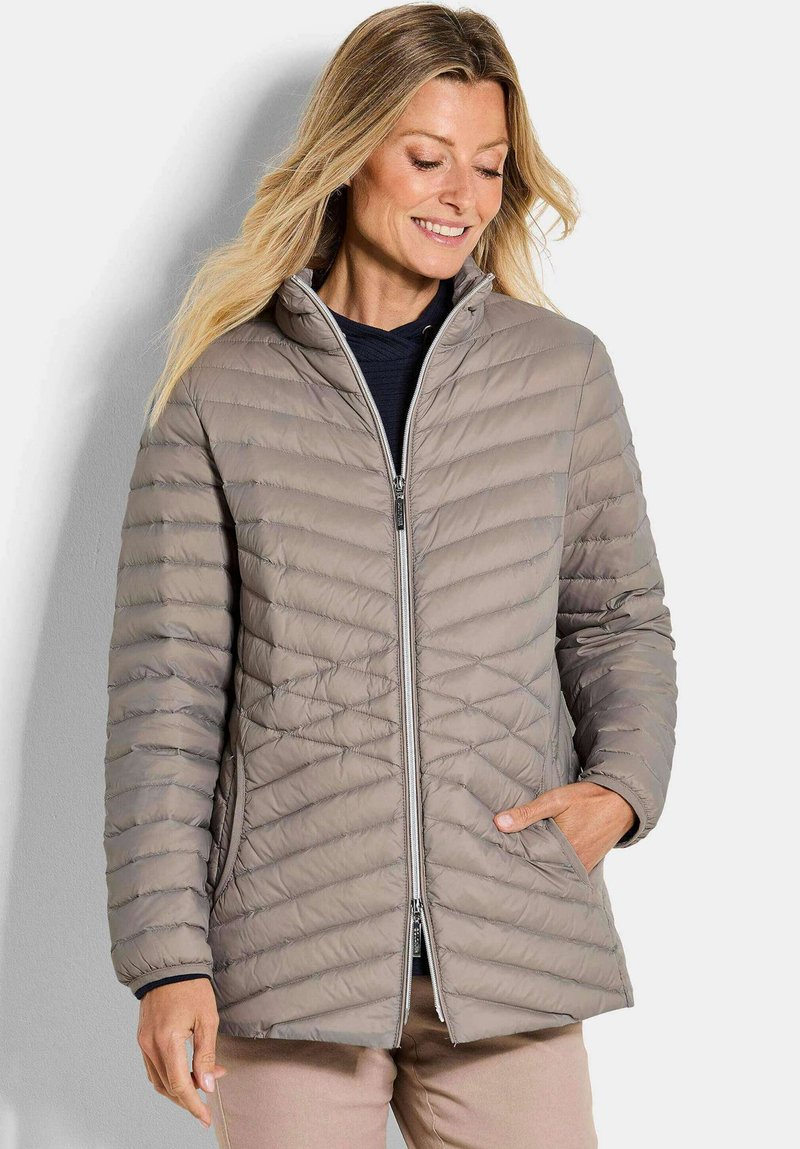 Vrouw in een lichtbruine gewatteerde jas met een ritssluiting aan de voorkant, handen in de zakken, lachend en naar beneden kijkend tegen een effen achtergrond.