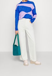 Person som bär en tröja med abstrakt mönster i blått och lila, vita vida byxor, beigefärgade klumpiga stövlar och håller en tealfärgad suède axelväska.