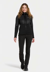 Melna džempera jaka ar rāvējslēdzēju, ar iesaiņotu augšdaļu, pieguļošām piedurknēm un divām kabatām ar rāvējslēdzējiem, kombinēta ar slaidām melnām biksēm un melniem zābakiem.