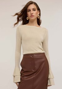 Top beige a maniche lunghe con texture a coste e scollo a barca, abbinato a una gonna a tubino in finta pelle marrone con tasca laterale e chiusura a bottone.