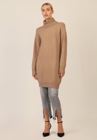 Strickrollkragenspullover in Hellbraun, oversized Schnitt, kombiniert mit grauen Jeans mit Fransensaum und schwarzen spitzen Absätzen.