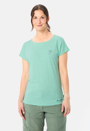 Jonge vrouw met bruin haar in een paardenstaart, die een muntgroen T-shirt met korte mouwen en een klein hartje draagt, en olijfgroene cargobroek.