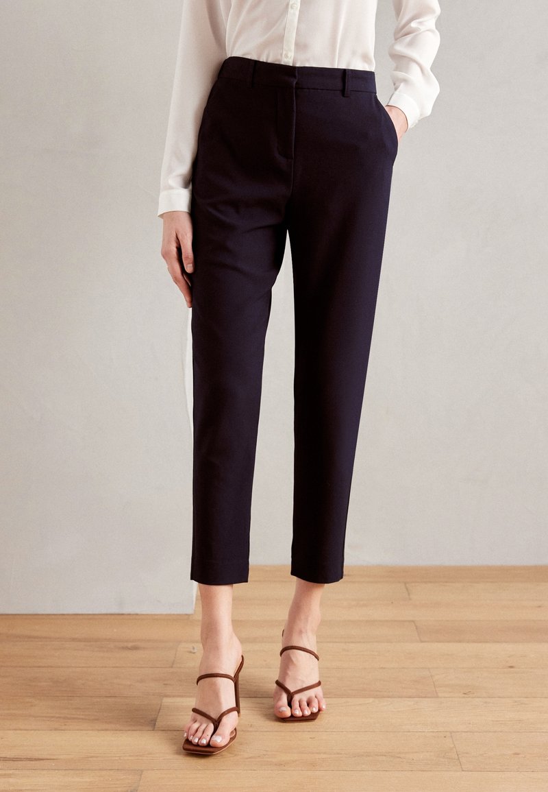 Femme portant un pantalon noir coupé sur mesure, une chemise blanche à manches longues et des sandales marron à talons, debout sur un sol en bois.