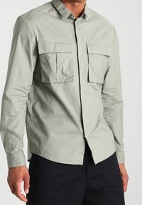 Mężczyzna w jasnoszarej koszuli zapinanej na guziki z dwiema dużymi kieszeniami na piersi i czarnych spodniach, stojący na jednolitym tle.