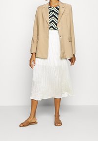 Blazer beige à deux boutons sur un haut à chevrons noir et blanc. Jupe midi plissée blanche et sandales à glisser beige complètent la tenue.