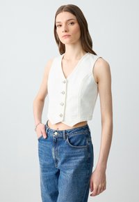 Gilet bianco corto con scollo a V, chiusura a cinque bottoni e orlo obliquo. Abbinato a jeans blu a vita alta, che mostrano un fit casual.