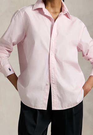 Persoon draagt een lichtroze blouse met knopen en een kraag, met lange mouwen en een zwarte broek, handen op de heupen rustend.