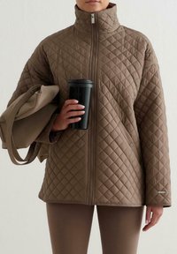 Brun quiltad jacka med hög krage och dragkedja framtill. Åtföljd av en beige väska och en svart rese-mugg, mot en neutral bakgrund.
