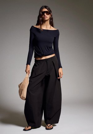 Vrouw die oversized zwarte pantalons met wijde pijpen draagt, een zwarte off-shoulder top, grote zonnebril, zwarte sandalen, een beige tas vasthoudend, staand tegen een grijze achtergrond.