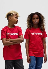 Röda crewneck-t-shirts med en vit "hummel"-logotyp, båda bärs av två barn med olika frisyrer, kombinerade med jeans.