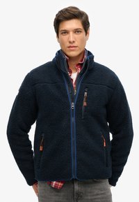 Marineblauwe fleece jas met een gestructureerde, pluizige buitenkant; heeft ritssluitingen in de zakken en oranje hardwareaccenten, een hoge kraag en blauwe afwerking.