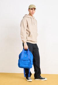 Modrý batoh s několika přihrádkami, vyrobený z odolné tkaniny. Nese ho osoba v béžové mikině s kapucí a černých kalhotách, stojící na žlutém povrchu.