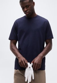 Άνδρας με μπλε ναυτικό πουκάμισο που κρατάει λευκό γάντι γκολφ Adidas μπροστά σε απλό φωτεινό φόντο.