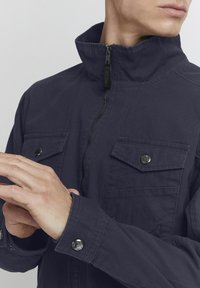 Mørkeblå zip-up jakke med høj krave, to brystlommer, sølvfarvede trykknapper og et tekstureret stof. Har en slank, skræddersyet pasform.