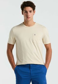 Beige T-shirt met korte mouwen, gemaakt van zacht materiaal, met een voorzak waarin een in marineblauw geborduurd pinguïnlogo is verwerkt.